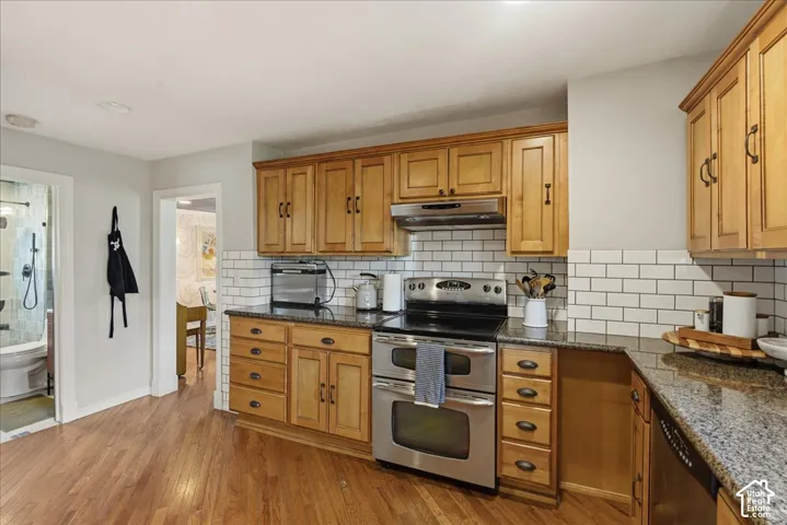 Wood-finish flooring, wood cabinetry with dark hardware, granite countertops, subway tile backsplash, and stainless steel appliances