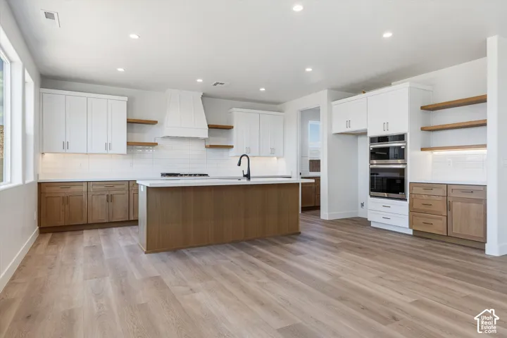Spacious kitchen featuring wood-finish flooring, white shaker cabinetry, an expansive island with a matte black faucet, integrated stainless steel double ovens, and open wood shelving