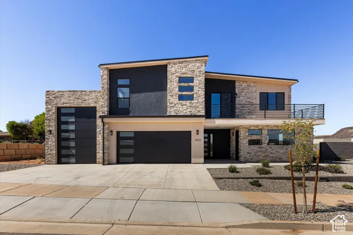 Modern architectural exterior featuring stacked stone masonry, contrasting dark smooth-finish siding, a second-story balcony with metal railing, and horizontal-panel garage doors