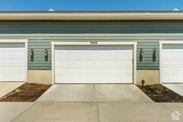 White paneled garage doors, exterior wall siding in a light green hue, contrasting white trim, exterior wall sconces, and concrete driveway