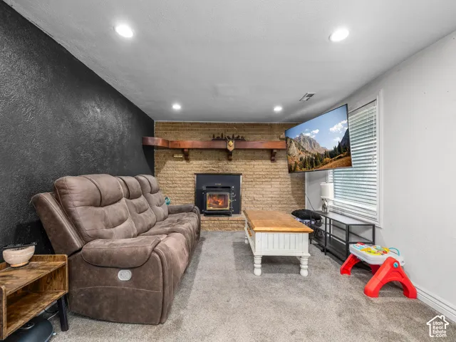 Carpeted flooring, brick fireplace surround with wood-burning stove, recessed lighting, and a prominent window with blinds
