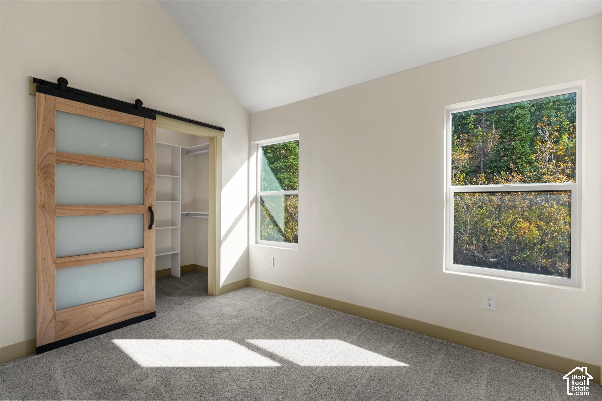 Vaulted ceiling room featuring a sliding barn door with frosted glass panels, built-in closet shelving, and two windows