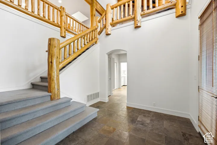 Stone tile flooring, natural wood log staircase railing, carpeted stairs, arched entryway, and a textured glass door with integrated blinds