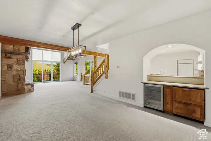 Vaulted ceiling with exposed wood beams, stone fireplace, floor-to-ceiling windows, wood staircase, and a built-in wet bar with cabinetry