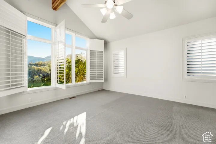 Vaulted ceiling with exposed wood beams, ceiling fan light fixture, expansive windows with plantation shutters, additional windows with plantation shutters, and light grey carpeting
