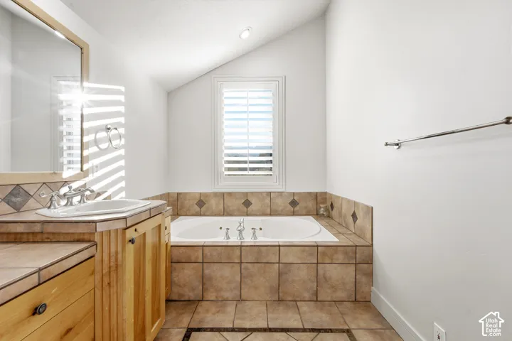 Built-in soaking tub with tile surround, wood vanity with integrated sink, recessed lighting, and a window with plantation shutters