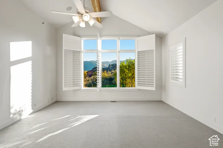 Vaulted ceiling with exposed wood beams, ceiling fan with integrated lighting, large picture windows with plantation shutters, additional window with plantation shutters, and light-toned carpeting
