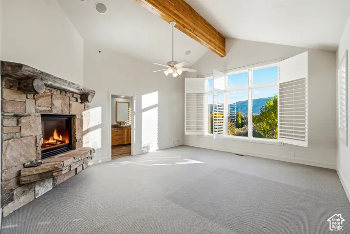 Vaulted ceiling with exposed wood beams, stone fireplace with hearth, large windows with interior shutters, ceiling fan, and carpeted flooring