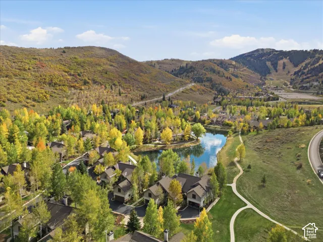 Residential properties surrounded by mature trees, a reflective pond, and a winding paved path, with mountainous terrain in the background