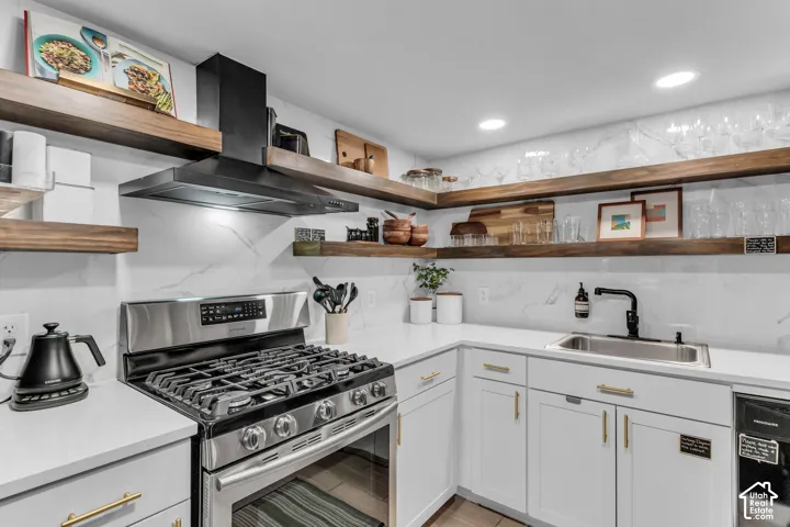 White cabinetry with gold-finish hardware, stainless steel gas range, black range hood, undermount sink with a black faucet, and open wood-finish shelving
