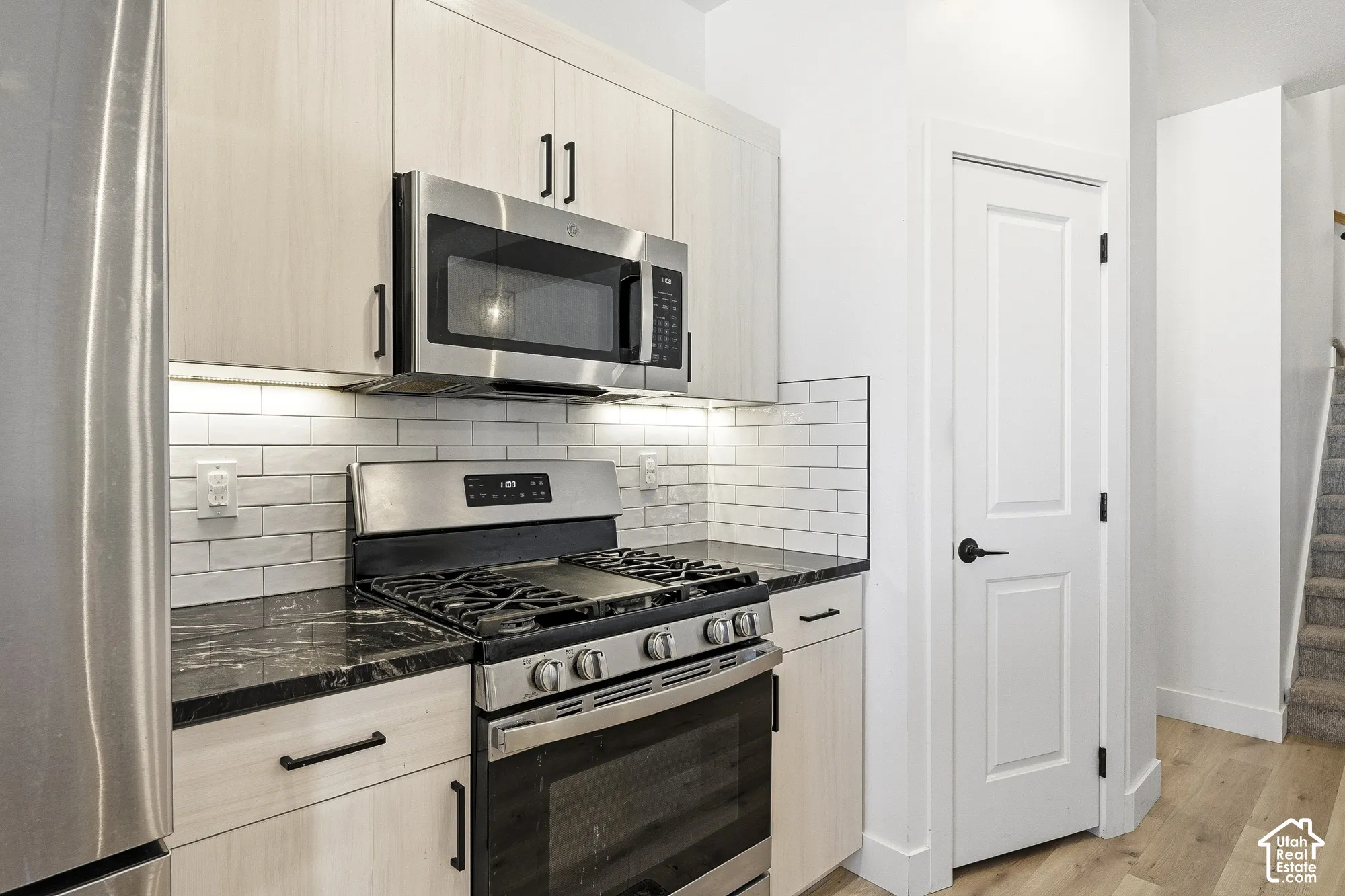 Stainless steel appliances, black countertops, light wood-finish cabinetry with black hardware, white subway tile backsplash, and light wood-finish flooring