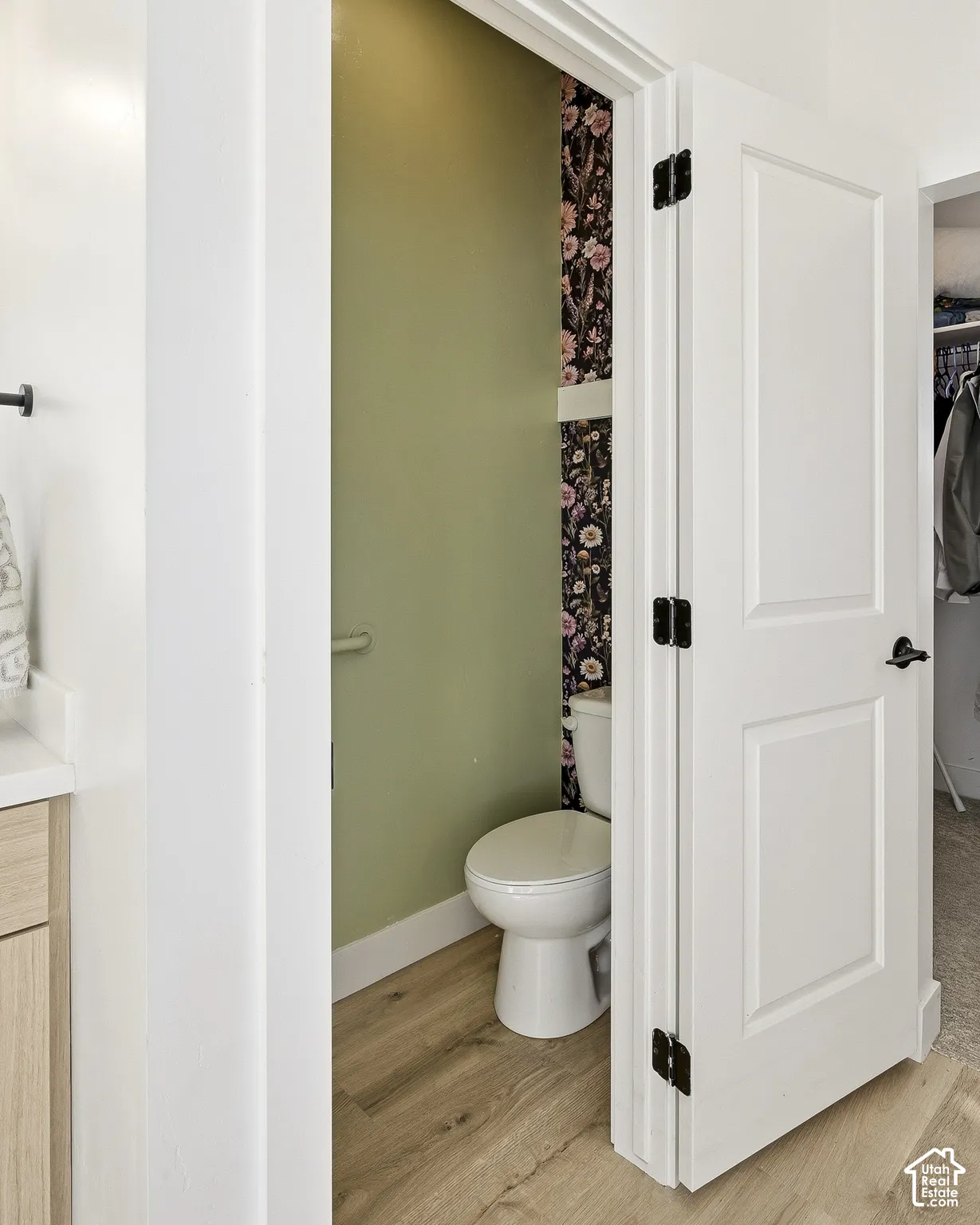 Light wood-finish flooring, white paneled door with matte black hardware, and toilet with a partial floral accent wall
