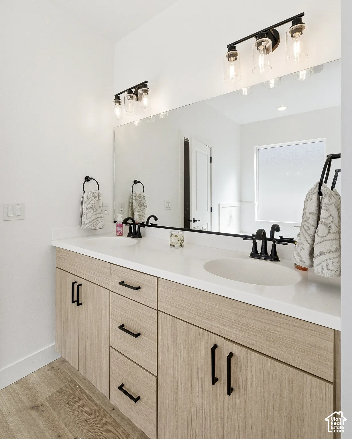 Dual vanity with integrated sinks, white countertops, wood-finish cabinetry, black hardware, and wood-finish flooring