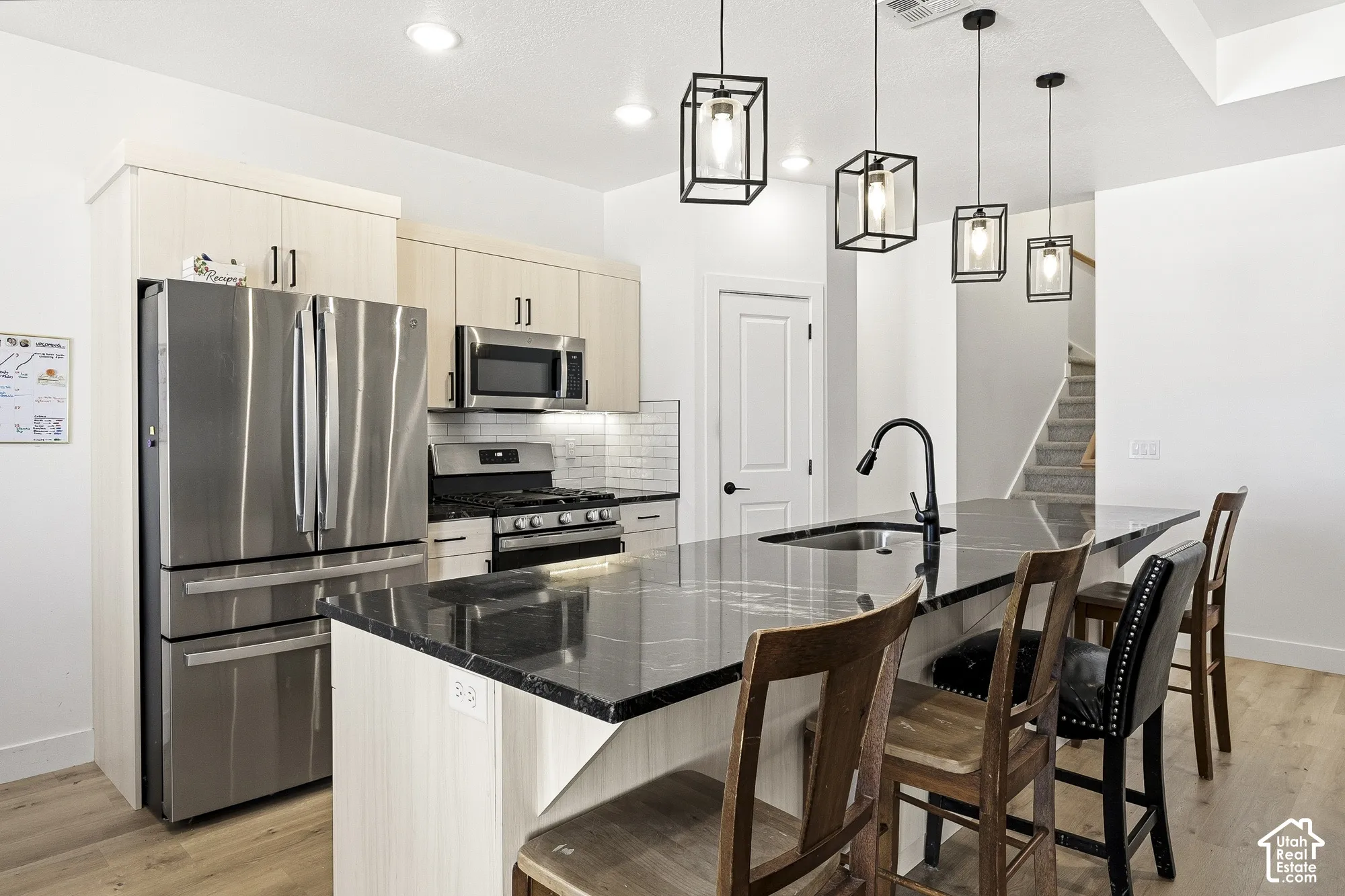 Kitchen featuring stainless steel appliances, light wood-finish cabinetry, a dark countertop island with an integrated sink, and modern pendant lighting