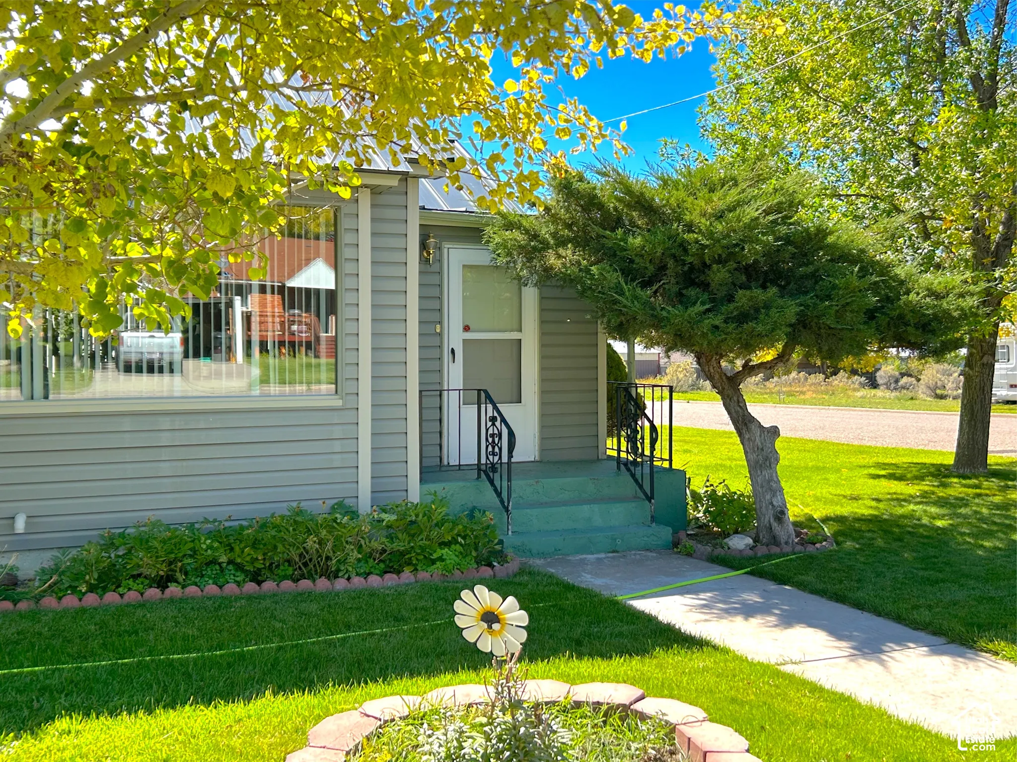 Charming residence featuring a manicured front lawn with mature shade trees, decorative planting beds with seasonal blooms, and a welcoming entrance with metal railings and concrete walkway