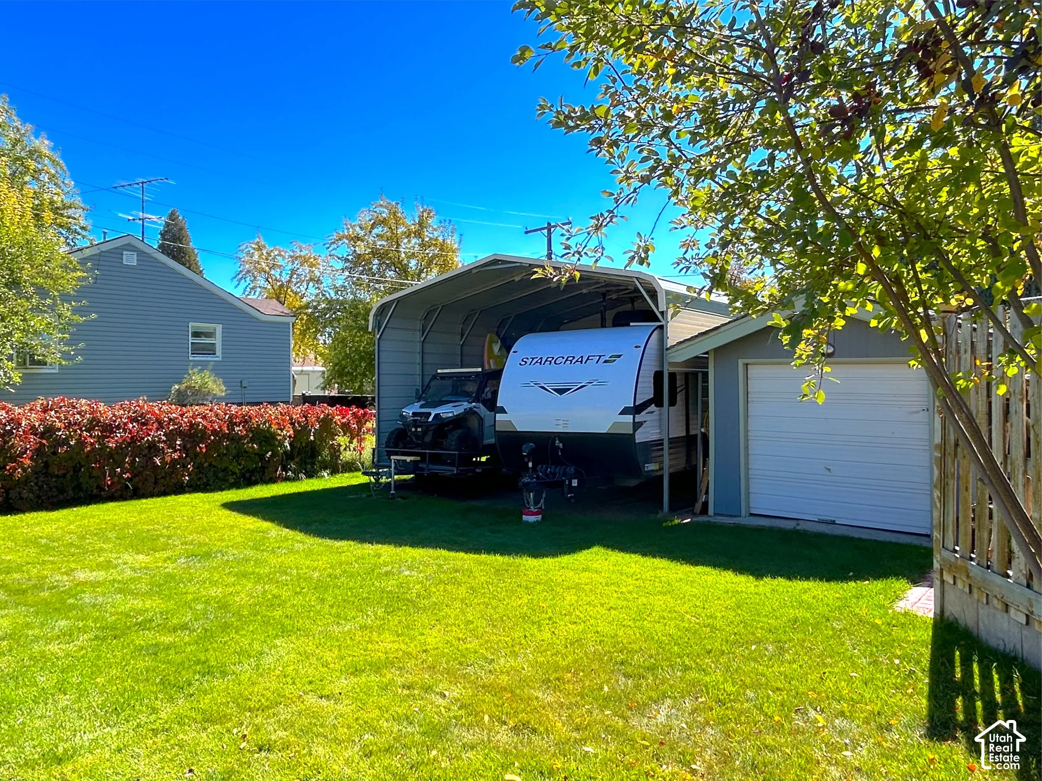 Expansive lawn area, mature perimeter landscaping, detached garage with roll-up door, metal carport structure, and a wood privacy fence