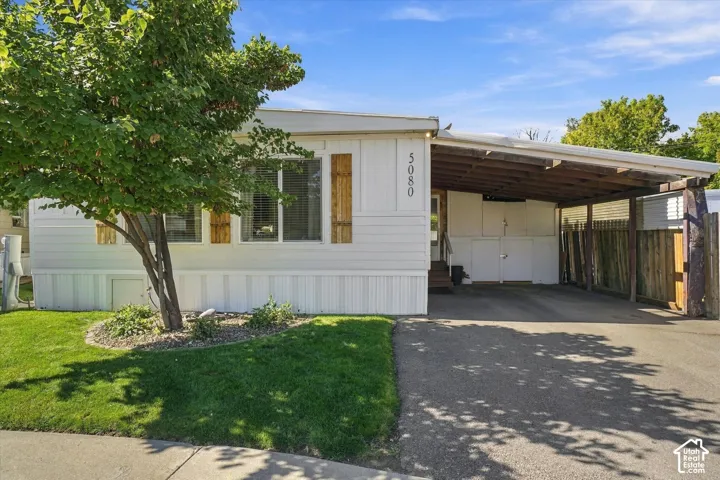 White siding exterior with a covered carport, wood-finish shutters, and a mature tree