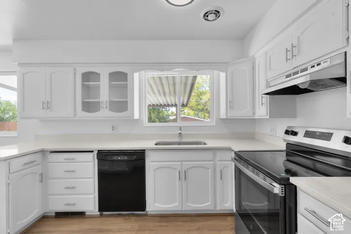 White cabinetry with glass-front accents, light-colored countertops, stainless steel sink, black built-in dishwasher, and a black range with stainless steel accents