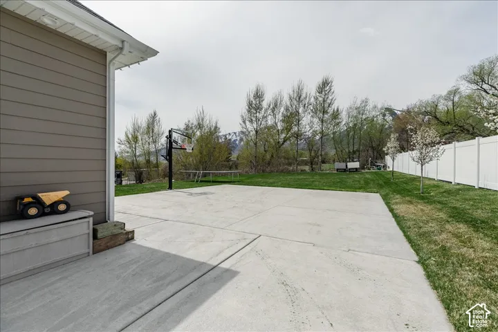 Spacious concrete pad, basketball hoop, lush green lawn, white privacy fencing, and distant mountain scenery
