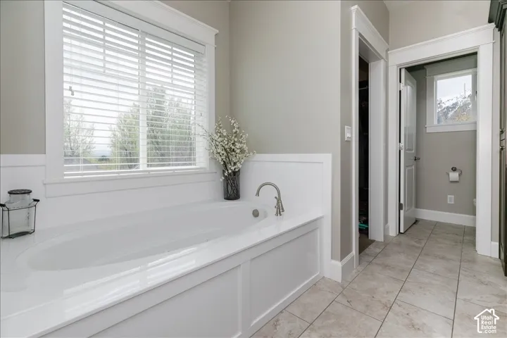 Built-in soaking tub with brushed nickel faucet, large window with blinds, light gray wall paint, white wainscoting, and tile flooring