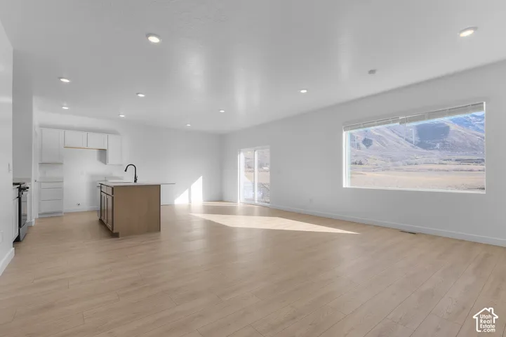 Open-concept living space with light wood-finish flooring, recessed lighting, a modern kitchen island with a black faucet, white cabinetry, and a large window framing distant mountains