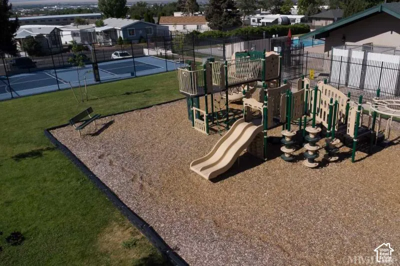 Recreational area featuring a playground structure with multiple slides, wood chip ground cover, a nearby bench, and fenced tennis courts