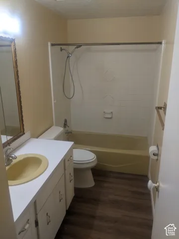 Wood-finish flooring, white vanity with cabinet storage, countertop with integrated sink, white tiled tub surround, and a chrome shower fixture