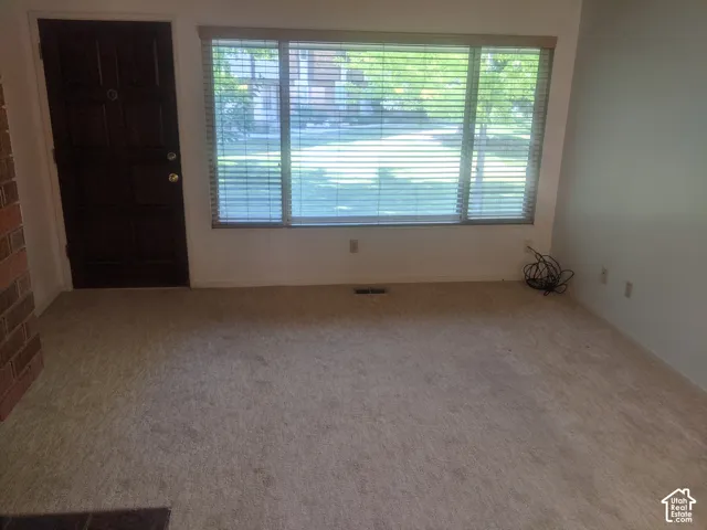 Carpeted flooring, white wall finishes, large window with horizontal blinds, dark panel door, and brick wall accent