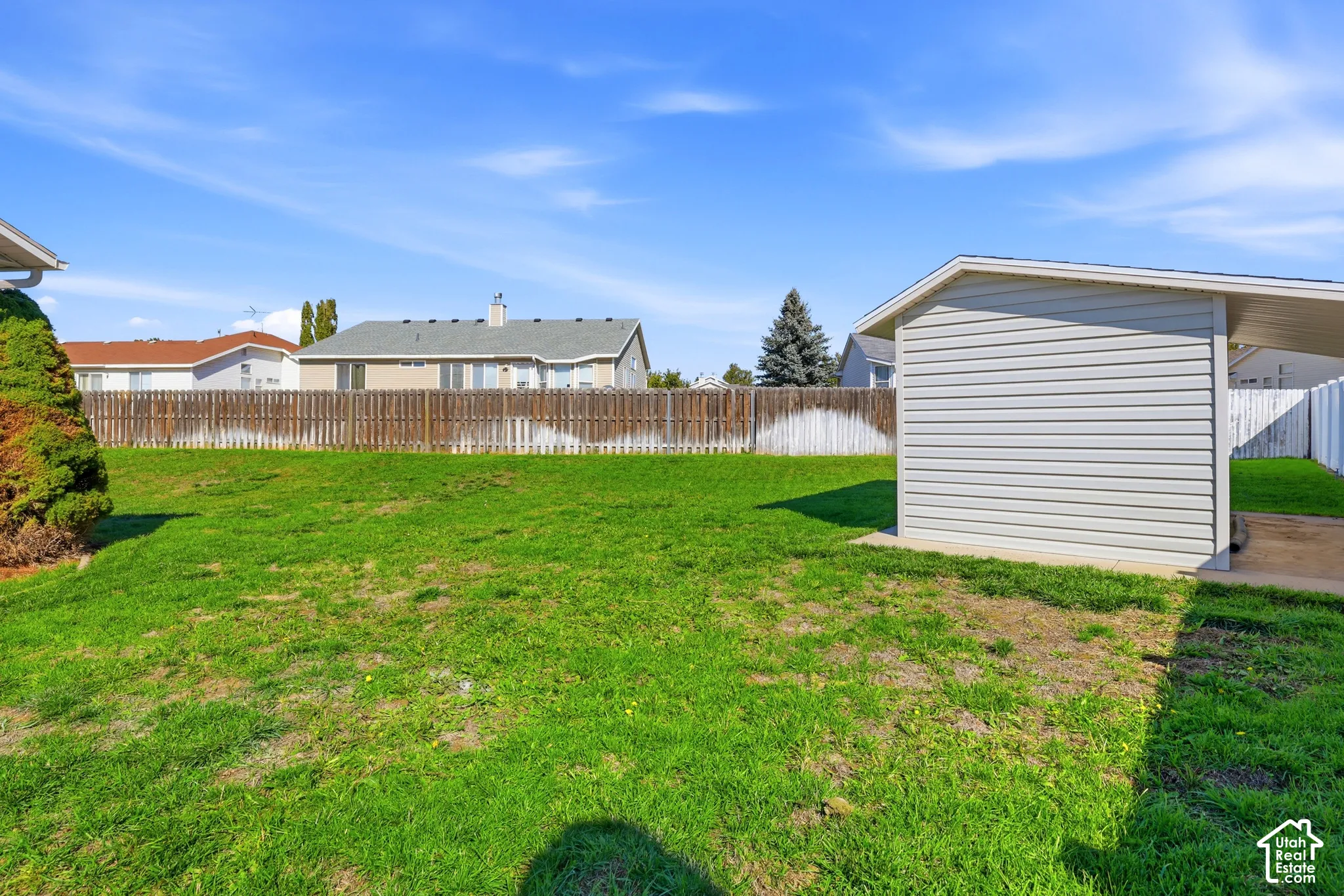 Expansive green space featuring a wood fence, mature landscaping, and a side-gabled carport with horizontal siding