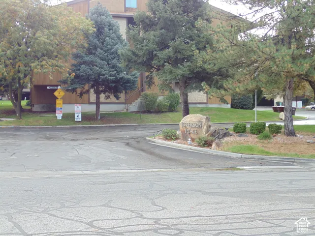 Community entrance featuring a stone monument sign, paved driveway, mature coniferous and deciduous trees, and landscaped grounds