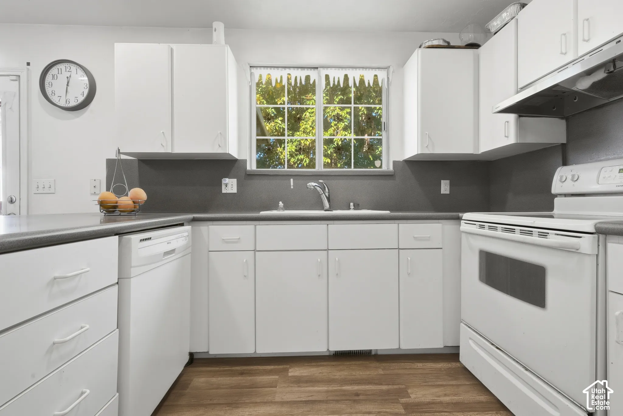 White cabinetry, wood-finish flooring, white built-in dishwasher, white electric range, and a stainless steel sink with a chrome faucet