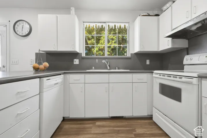 White cabinetry, wood-finish flooring, white built-in dishwasher, white electric range, and a stainless steel sink with a chrome faucet