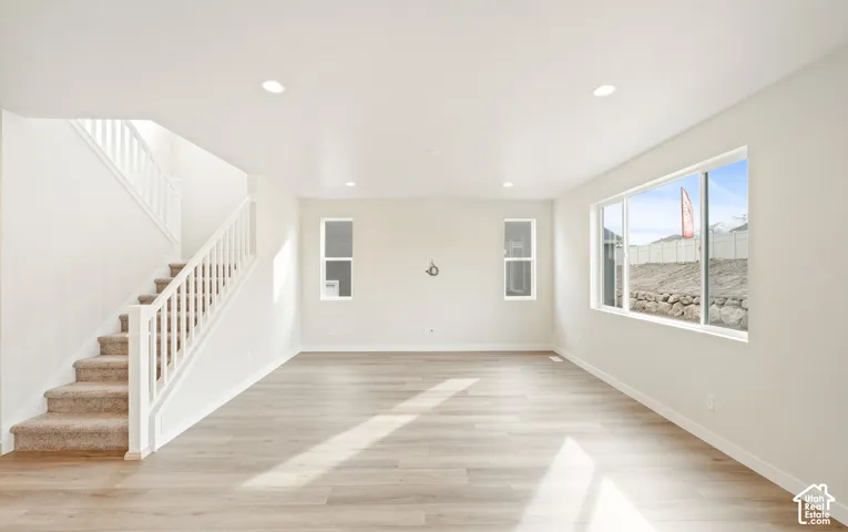 Light-toned wood-finish flooring, recessed ceiling lighting, a white painted staircase with carpeted treads, and large windows
