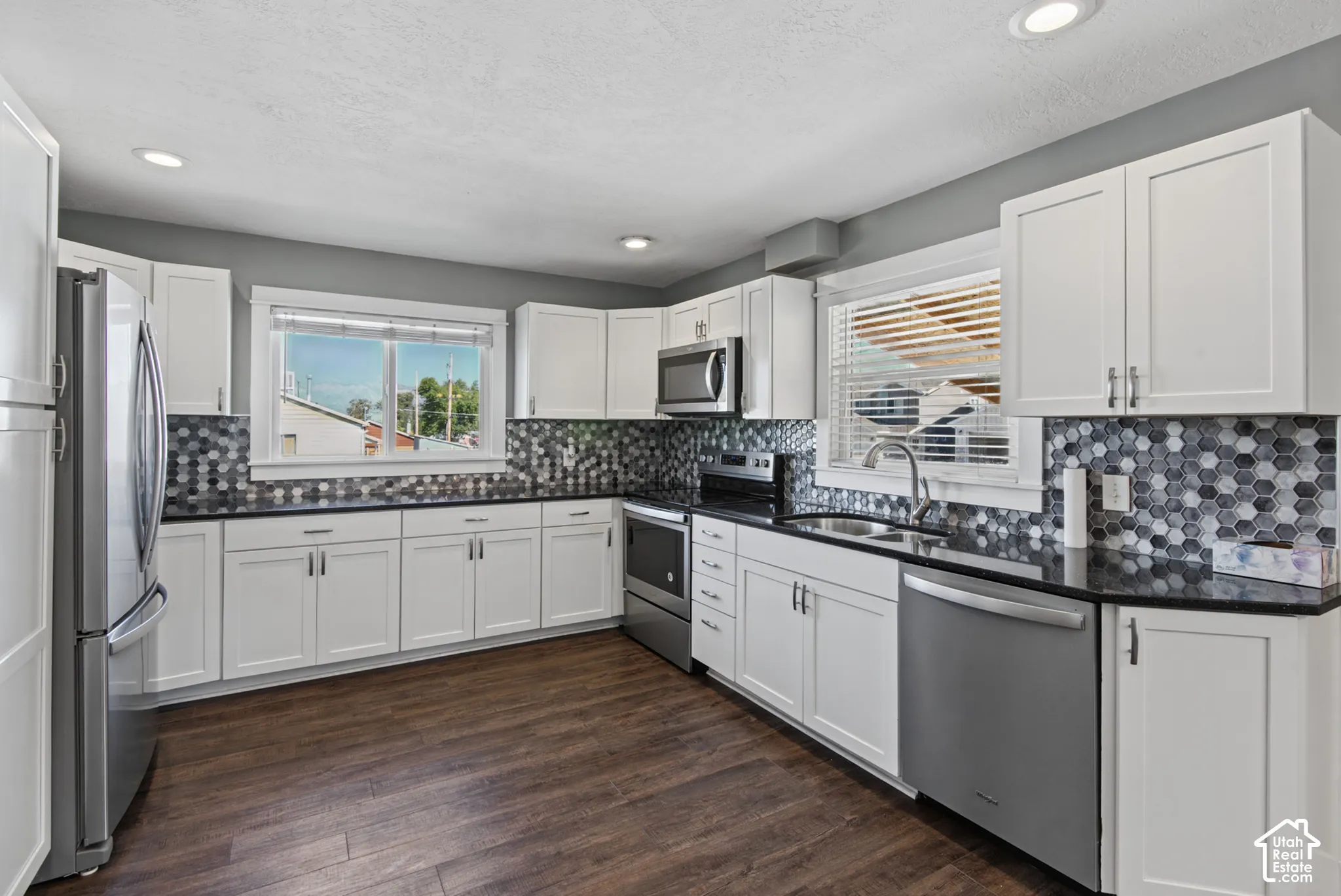 White cabinetry, dark countertops, a multi-tone hexagonal tile backsplash, stainless steel appliances, and wood-finish flooring