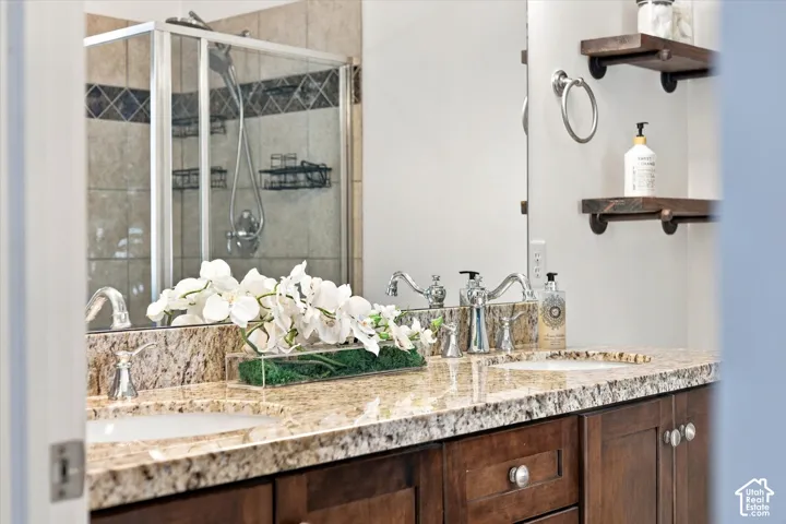 Double vanity with stone-surfaced countertops, undermount sinks, wood cabinetry, wall-mounted shelving, and a tiled shower enclosure