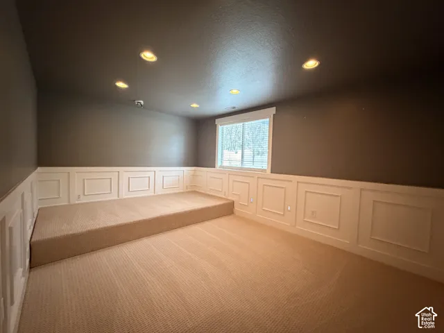 Recessed ceiling lighting illuminates a room featuring wainscoting, a raised carpeted platform, and a window with horizontal blinds