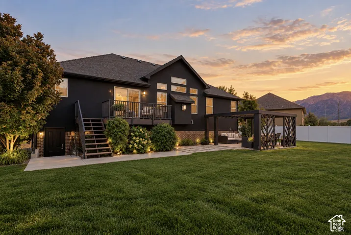Expansive rear yard featuring a manicured lawn, dark-toned exterior, elevated deck with staircase, modern pergola, and integrated exterior lighting
