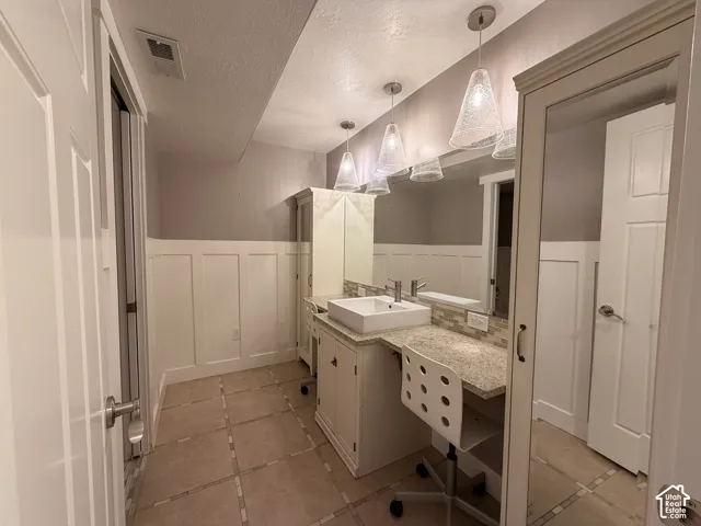 White wainscoting paneling, tile flooring, vanity with vessel sink, wall-mounted mirror, and pendant lighting