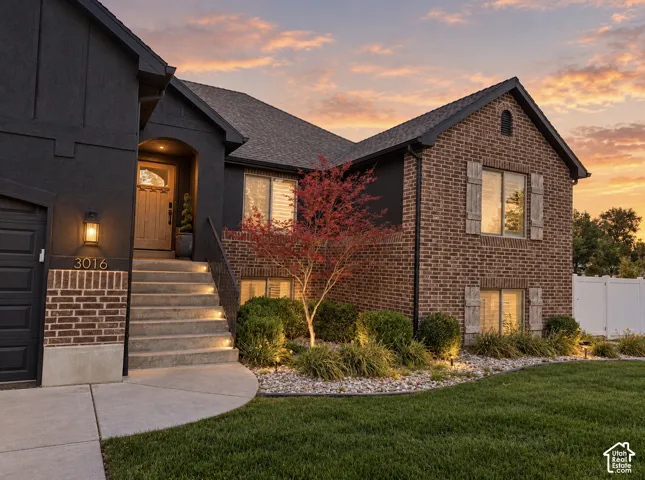 Brick and stucco exterior, prominent entry steps with integrated lighting, wood-finish front door, well-maintained landscaping with accent lighting, and a white privacy fence