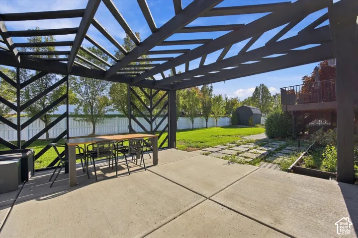 Custom geometric-patterned pergola over a concrete patio, stone paver pathway, manicured lawn, and perimeter white vinyl fencing