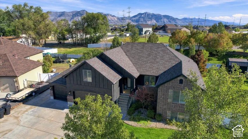 Multi-level exterior featuring a combination of brick and vertical metal siding, shingle roof, attached multi-car garage, and concrete driveway, set against a mountain backdrop