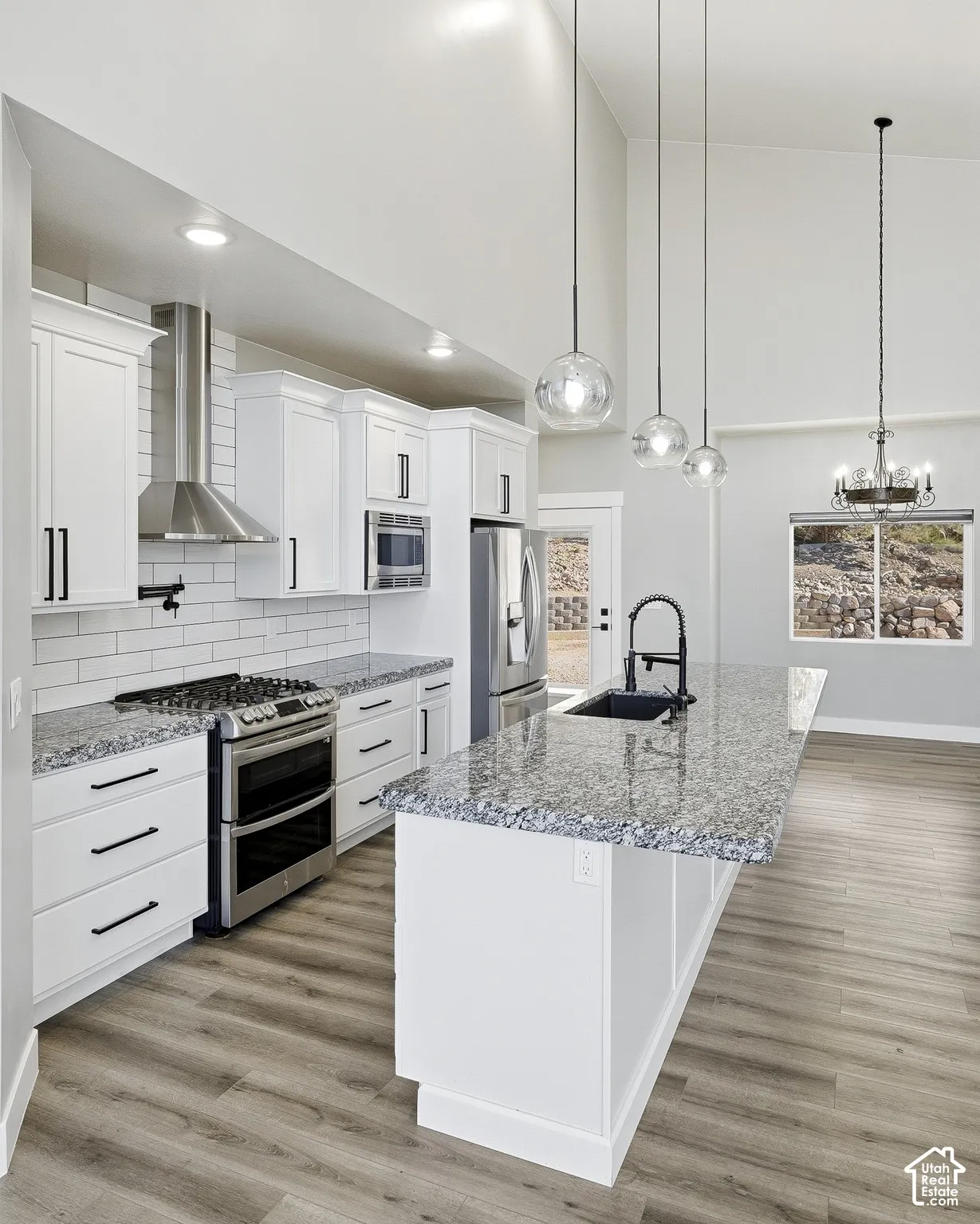 Vaulted ceiling with pendant lighting, white cabinetry with black hardware, stainless steel appliances, speckled stone countertops, and wood-finish flooring