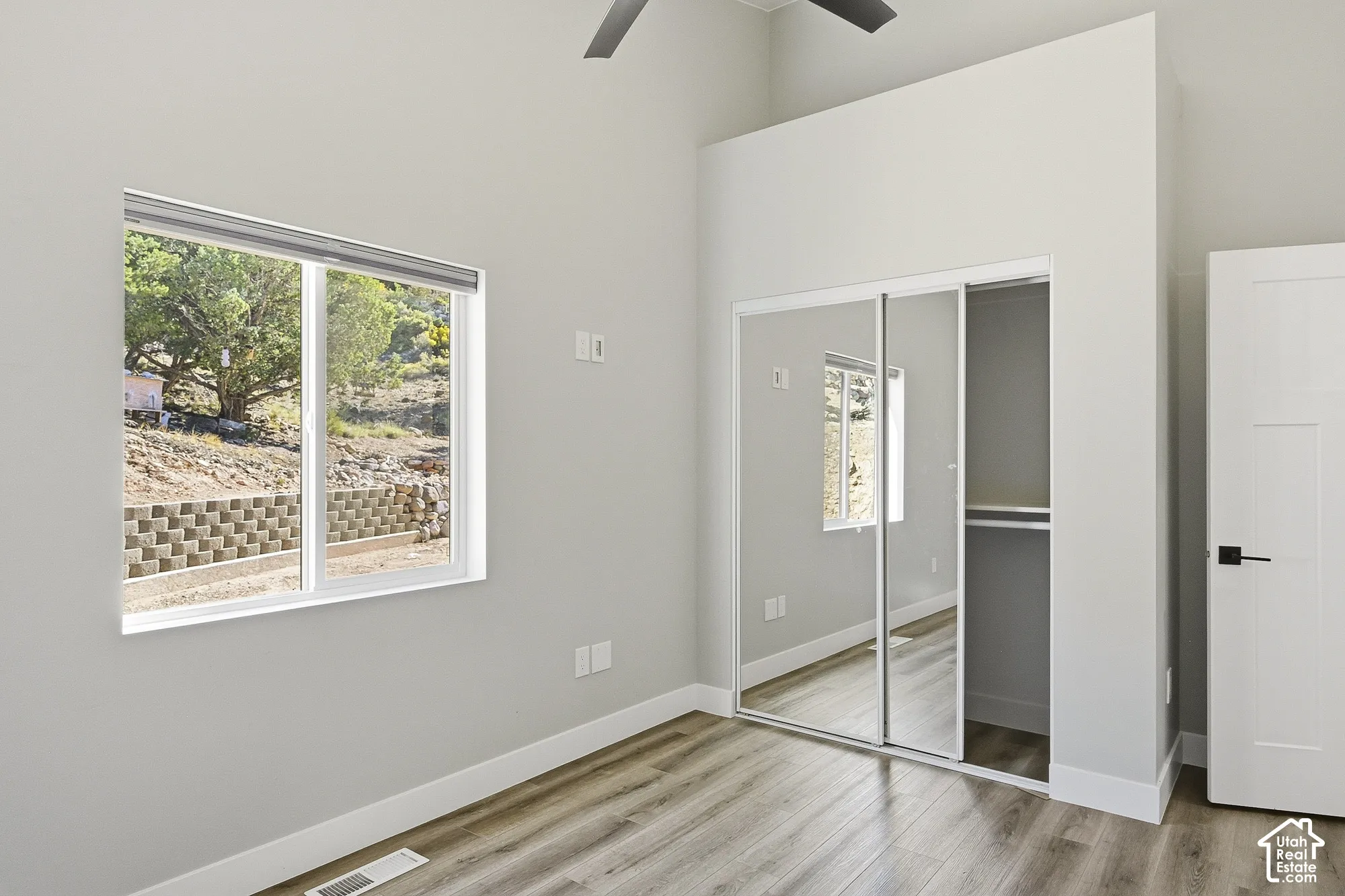 Window with exterior landscaping, wood-finish flooring, mirrored closet doors, built-in closet shelving, and a modern ceiling fan
