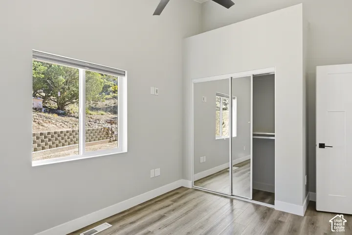 Window with exterior landscaping, wood-finish flooring, mirrored closet doors, built-in closet shelving, and a modern ceiling fan