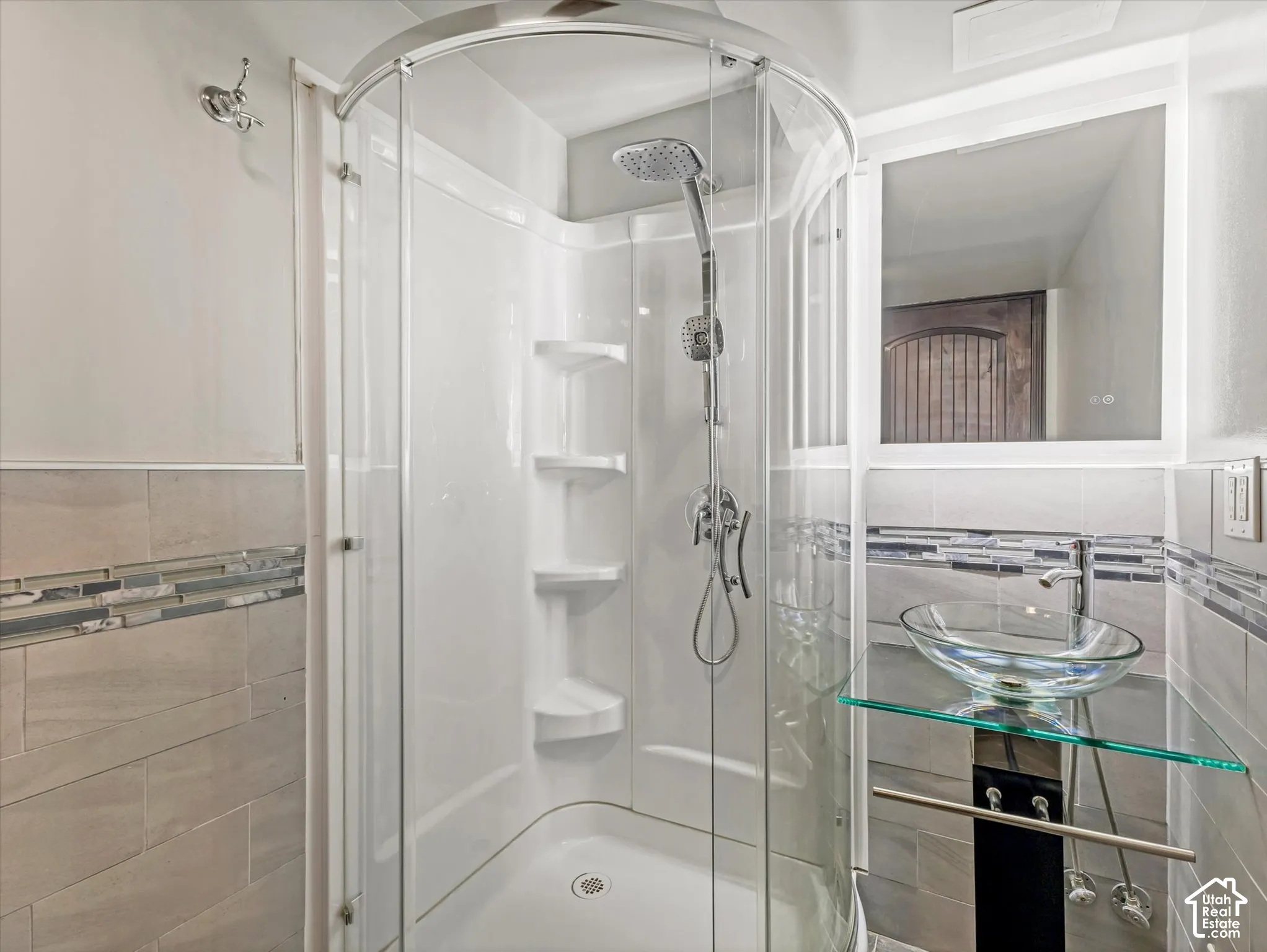 Curved glass shower enclosure with integrated shelving, chrome shower fixtures, glass vessel sink, glass vanity shelf, and tiled walls with decorative mosaic accent