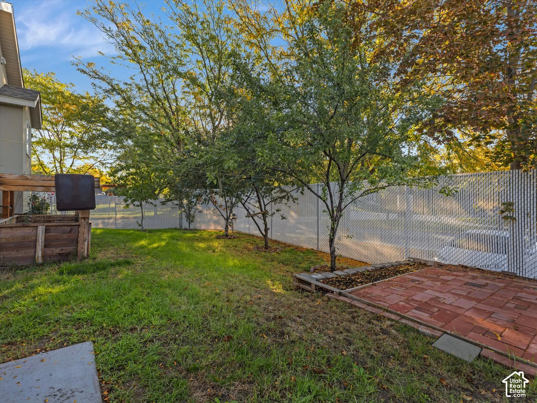 Grassed yard area featuring mature trees, a chain link fence, and a brick paver patio