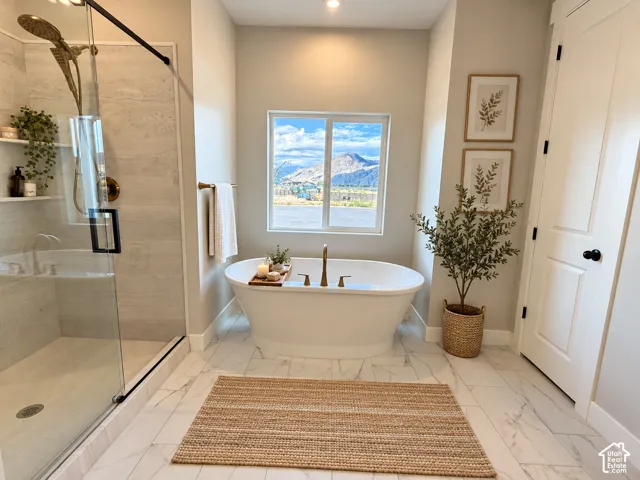 Freestanding soaking tub with a floor-mounted faucet, glass-enclosed shower with built-in shelving, large window showcasing mountain scenery, and white tile flooring