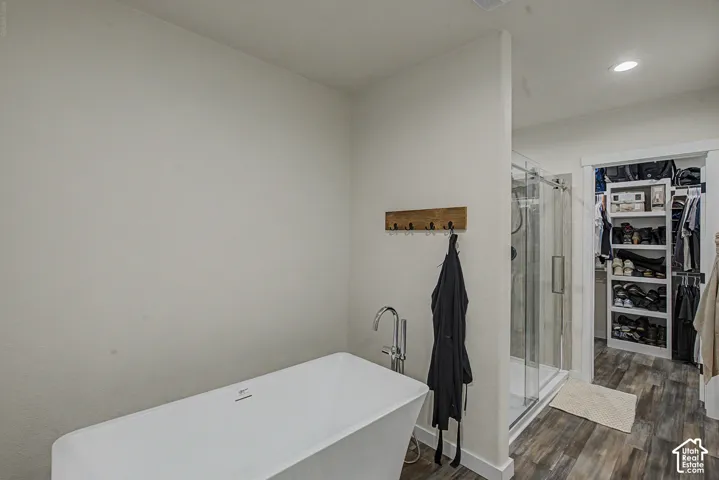 Freestanding soaking tub with floor-mounted faucet, glass-enclosed shower, wood-finish flooring, and recessed lighting