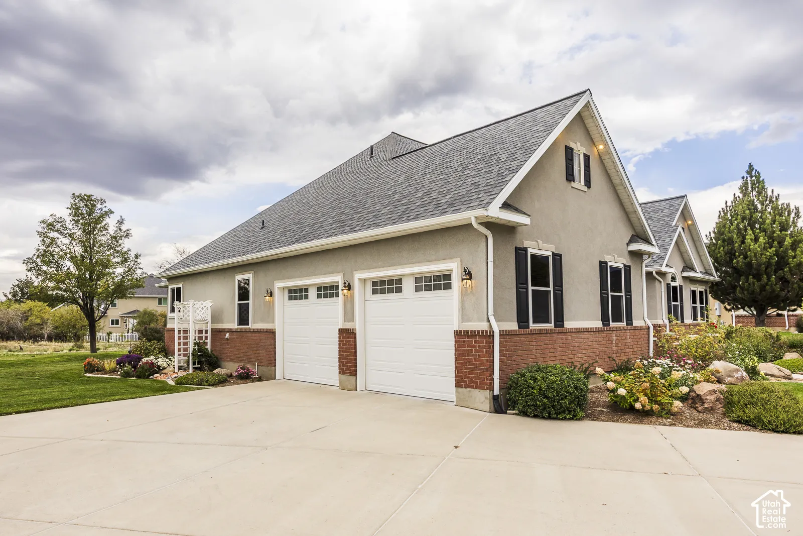 Stucco exterior with brick detailing, asphalt shingle roof, two-car garage with carriage-style doors, and established landscaping