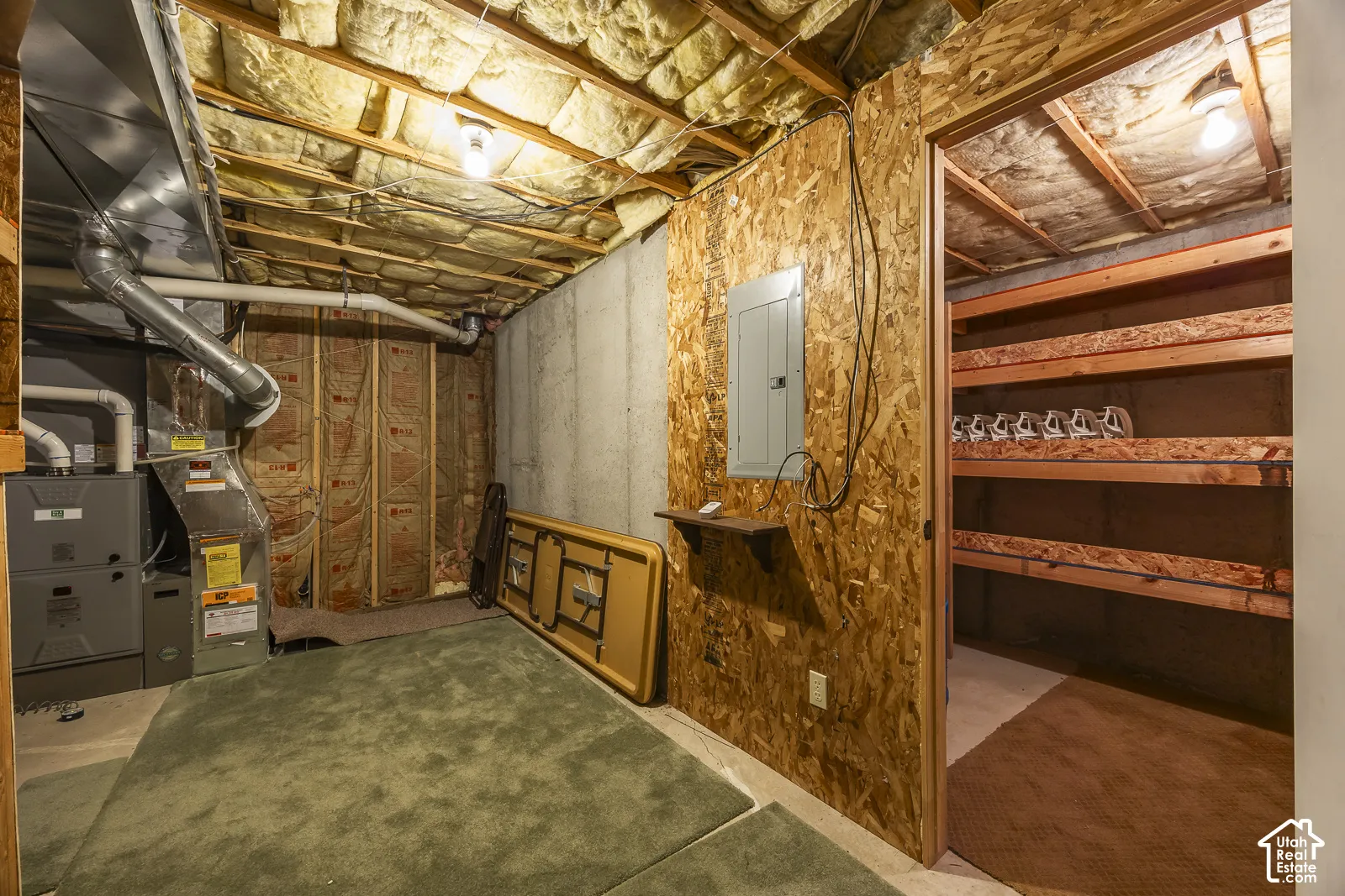 Unfinished basement space featuring a utility furnace, exposed ceiling joists with insulation, a concrete foundation wall, and an electrical panel